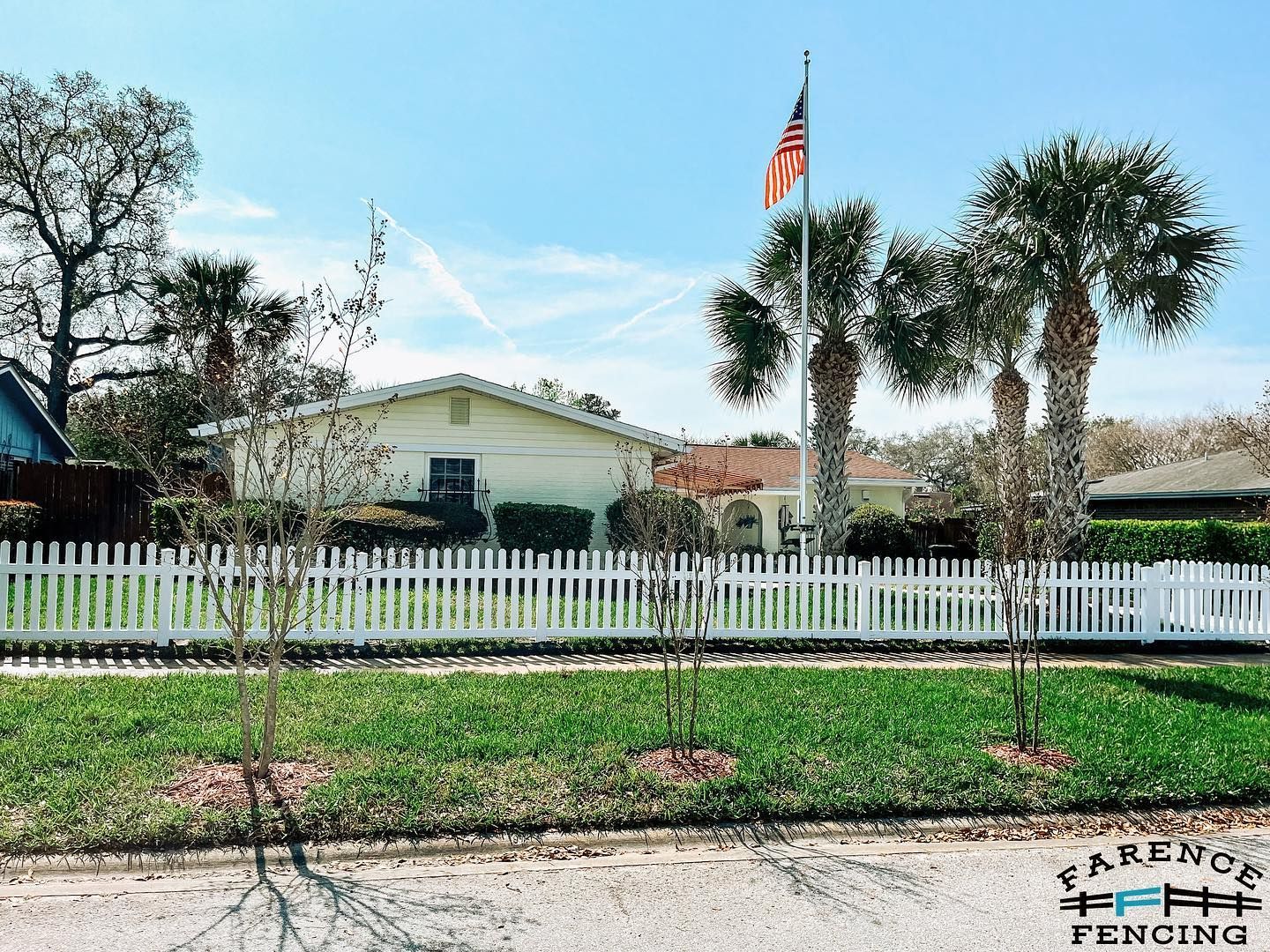 A white picket fence surrounds a house with palm trees in front of it.