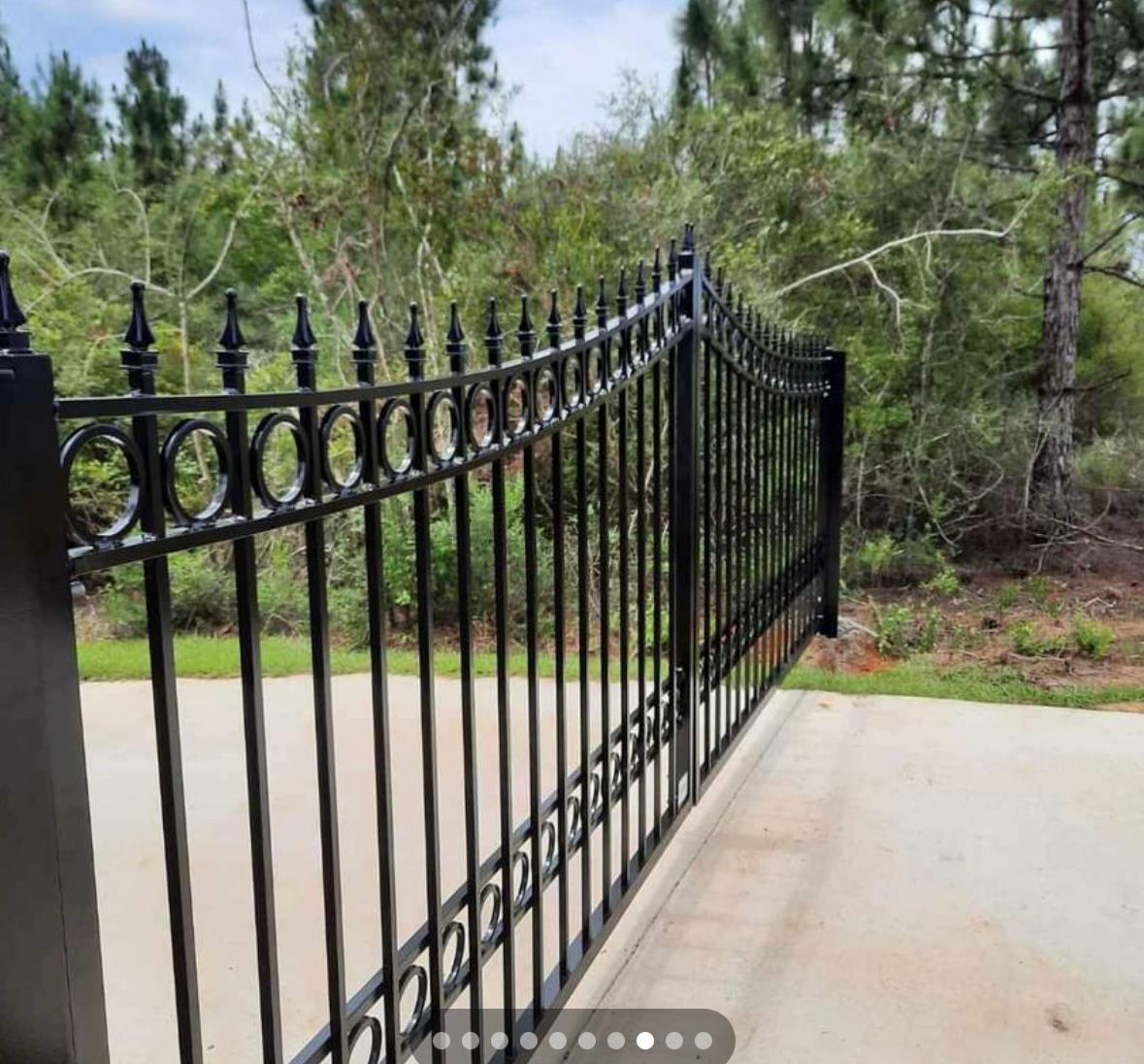 A black wrought iron gate is open to a driveway with trees in the background.