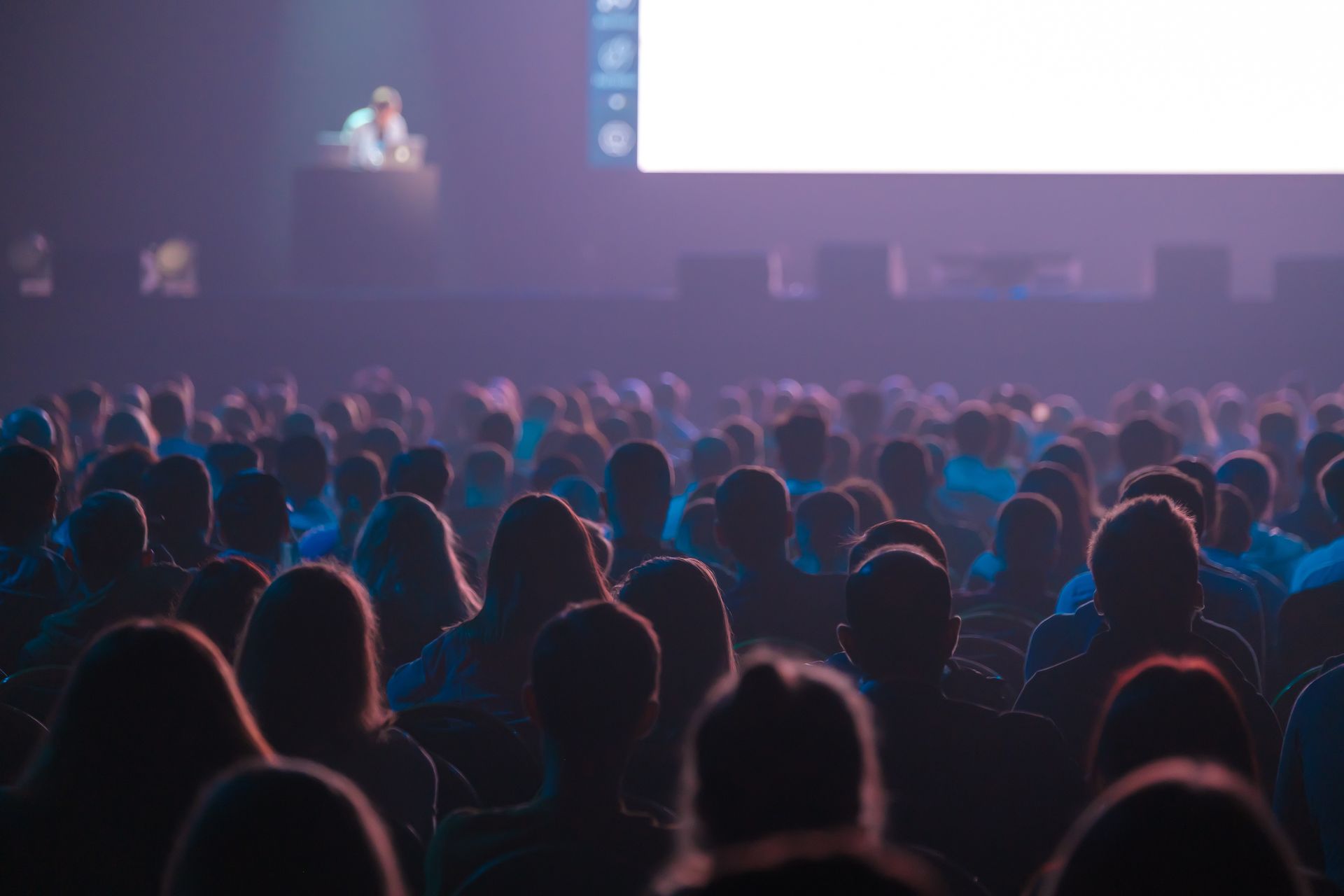 Audience in a dim room watches a presentation on a screen. Red chairs, windows, and dark decor.