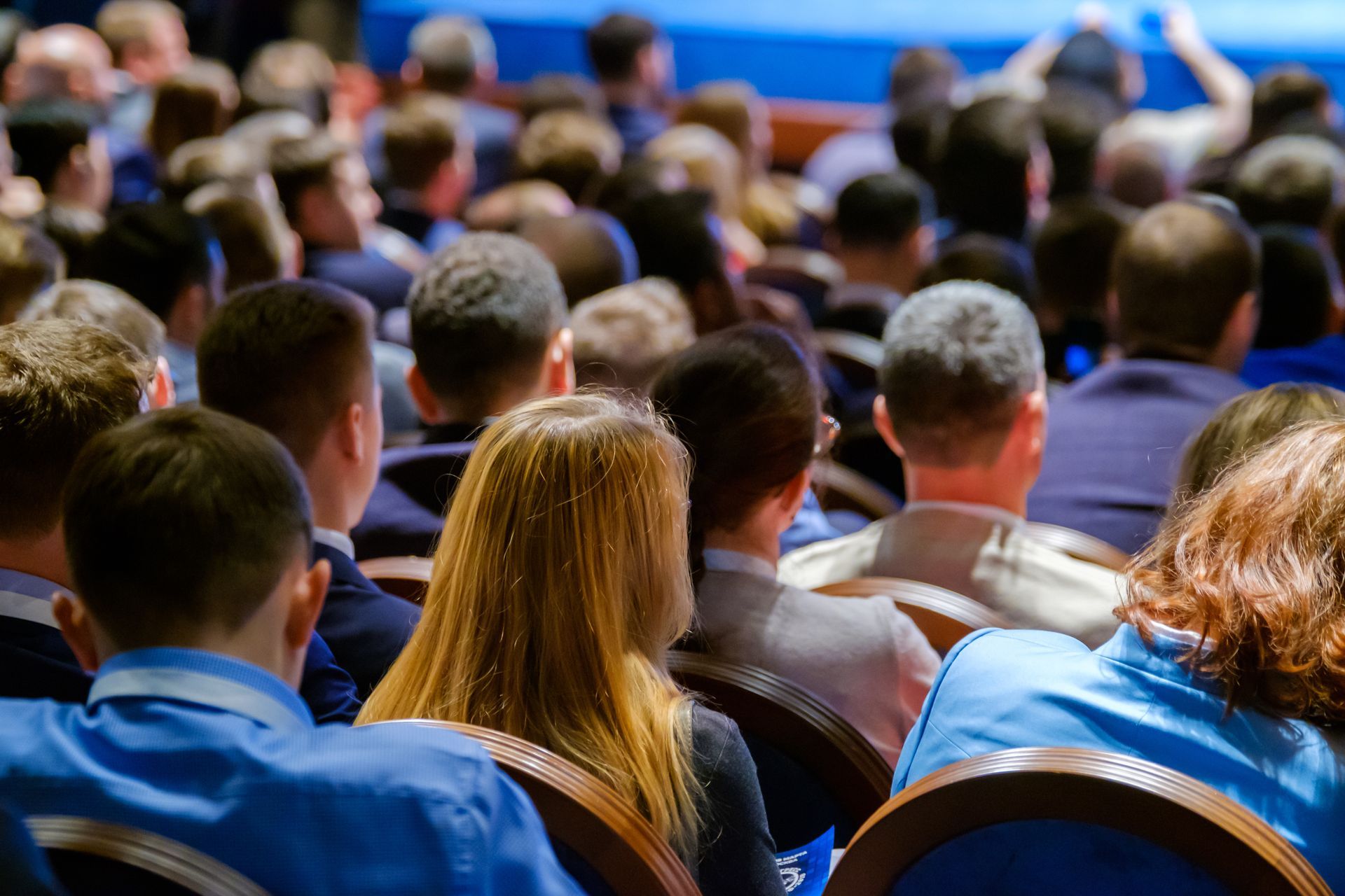 Audience seated in rows, facing forward at a conference or seminar. Many people, varied hair colors, some wearing suits.