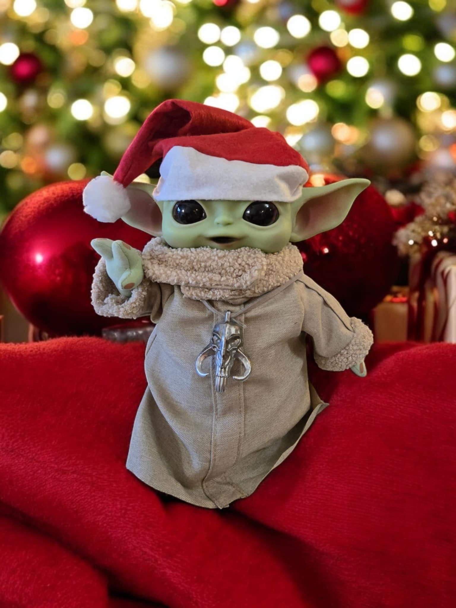 A baby yoda wearing a santa hat is sitting on a red blanket in front of a christmas tree.