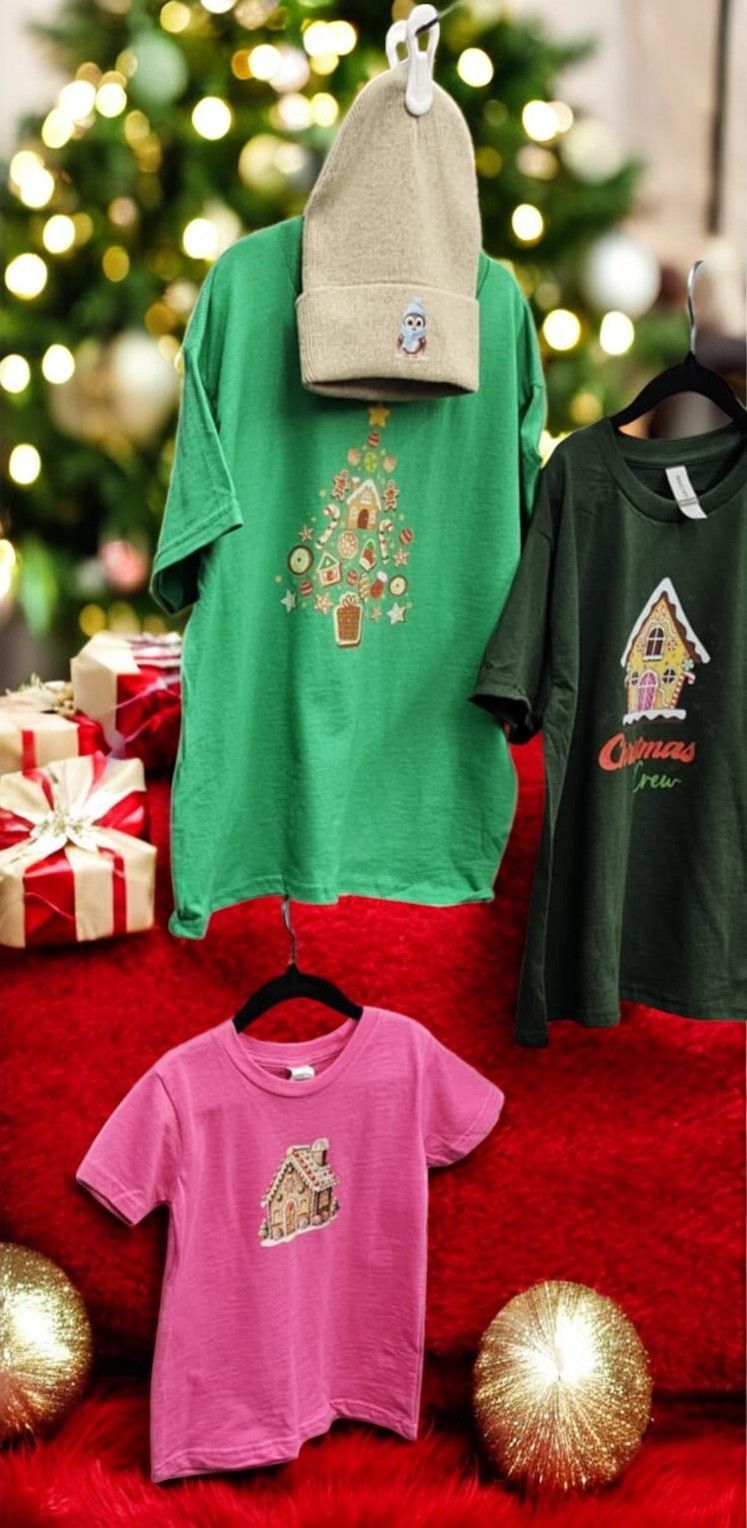 A green t-shirt with a christmas tree on it is hanging on a hanger next to a pink t-shirt.