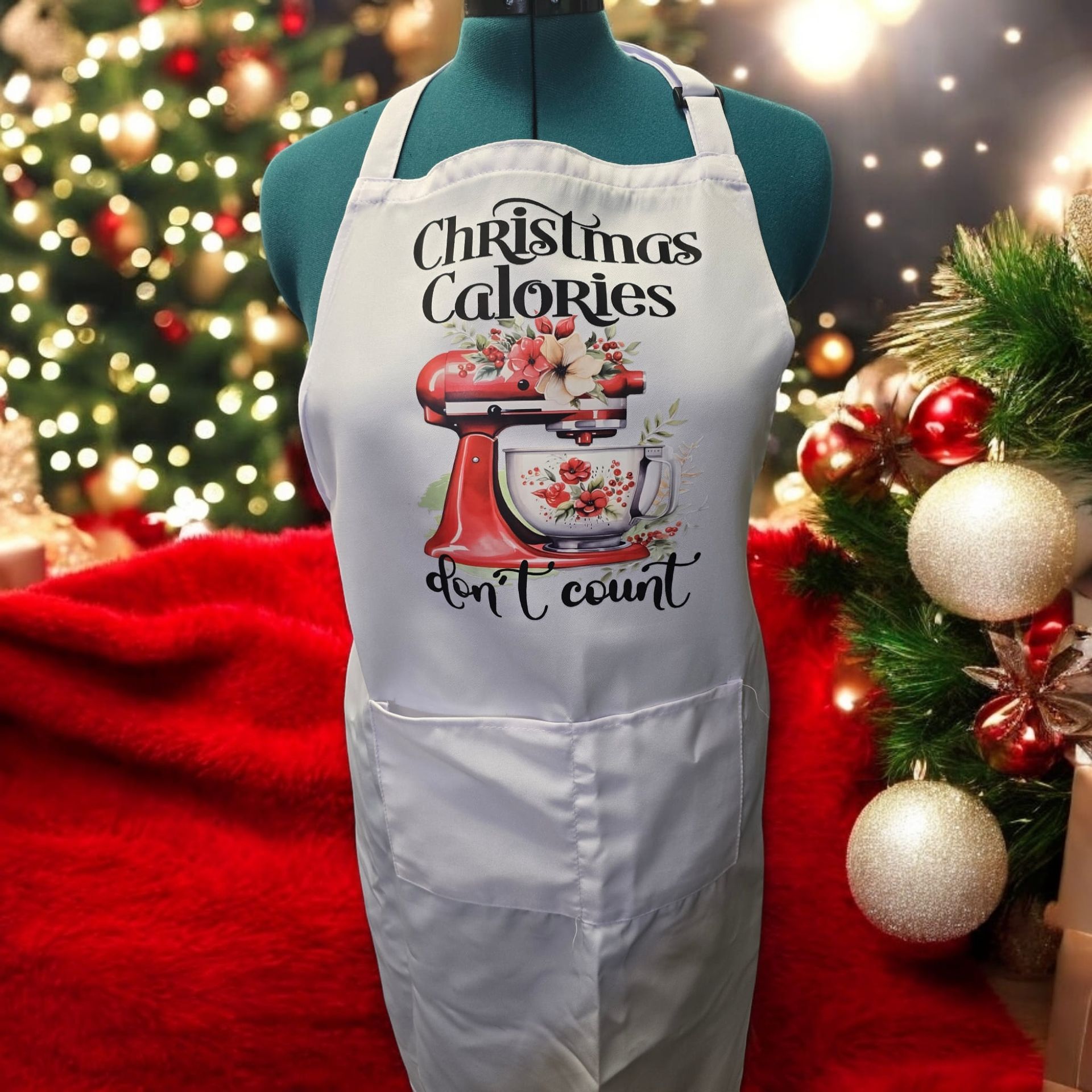 A white apron that says christmas calories don 't count is on a mannequin in front of a christmas tree.