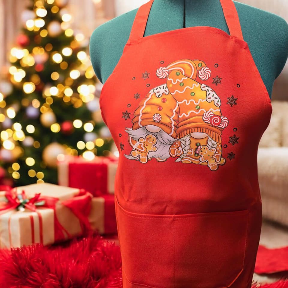 A red apron with a gingerbread gnome on it is on a mannequin in front of a christmas tree.