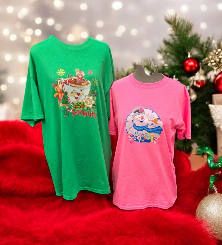 A green t-shirt and a pink t-shirt are sitting on a red blanket in front of a christmas tree.