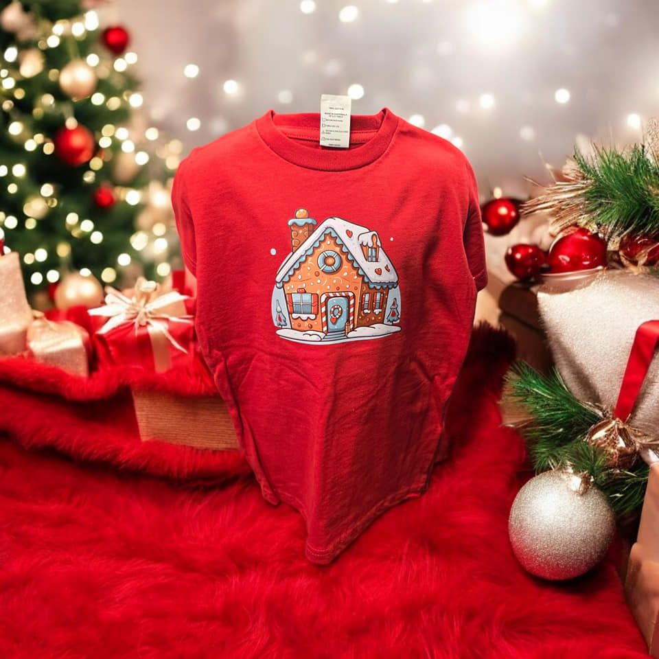 A red t-shirt with a gingerbread house on it is sitting on a red blanket next to a christmas tree.