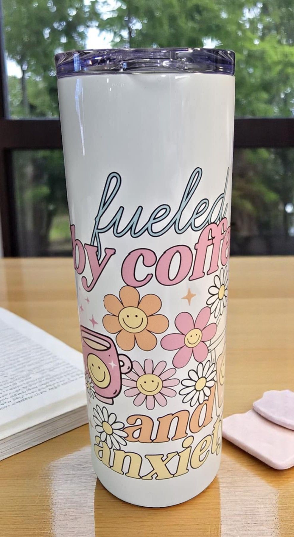 A stainless steel tumbler with flowers on it is sitting on a wooden table next to a book.