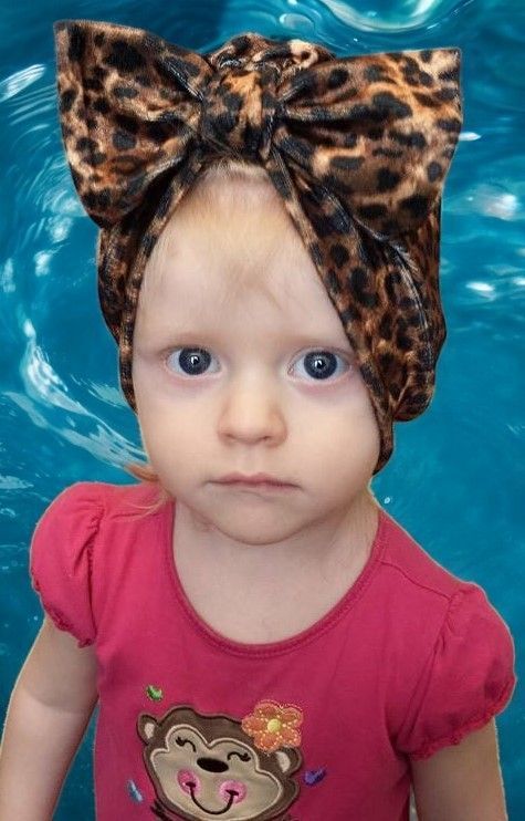 A little girl wearing a leopard print turban is standing in front of a pool.