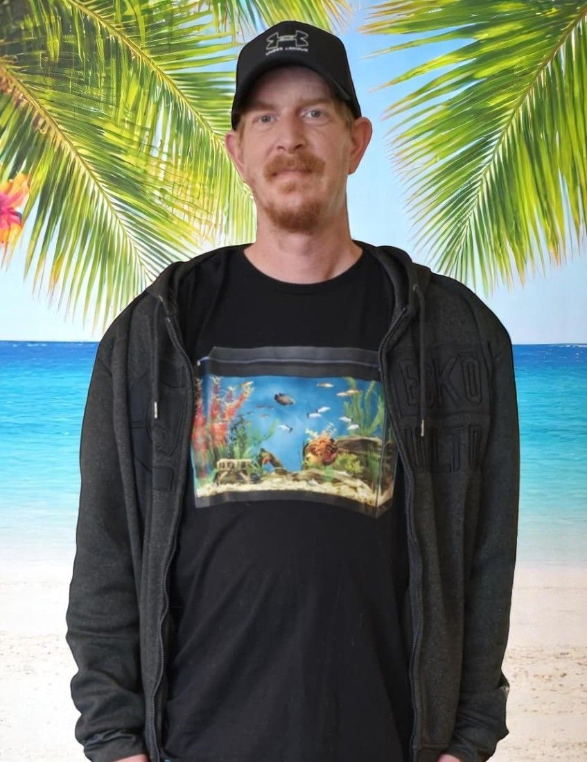 A man wearing a black shirt with an aquarium on it is standing on a beach.