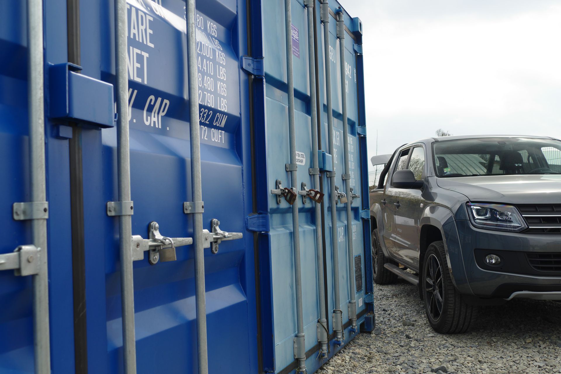 A grey pickup truck parked next to a large blue shipping container on a gravel surface.