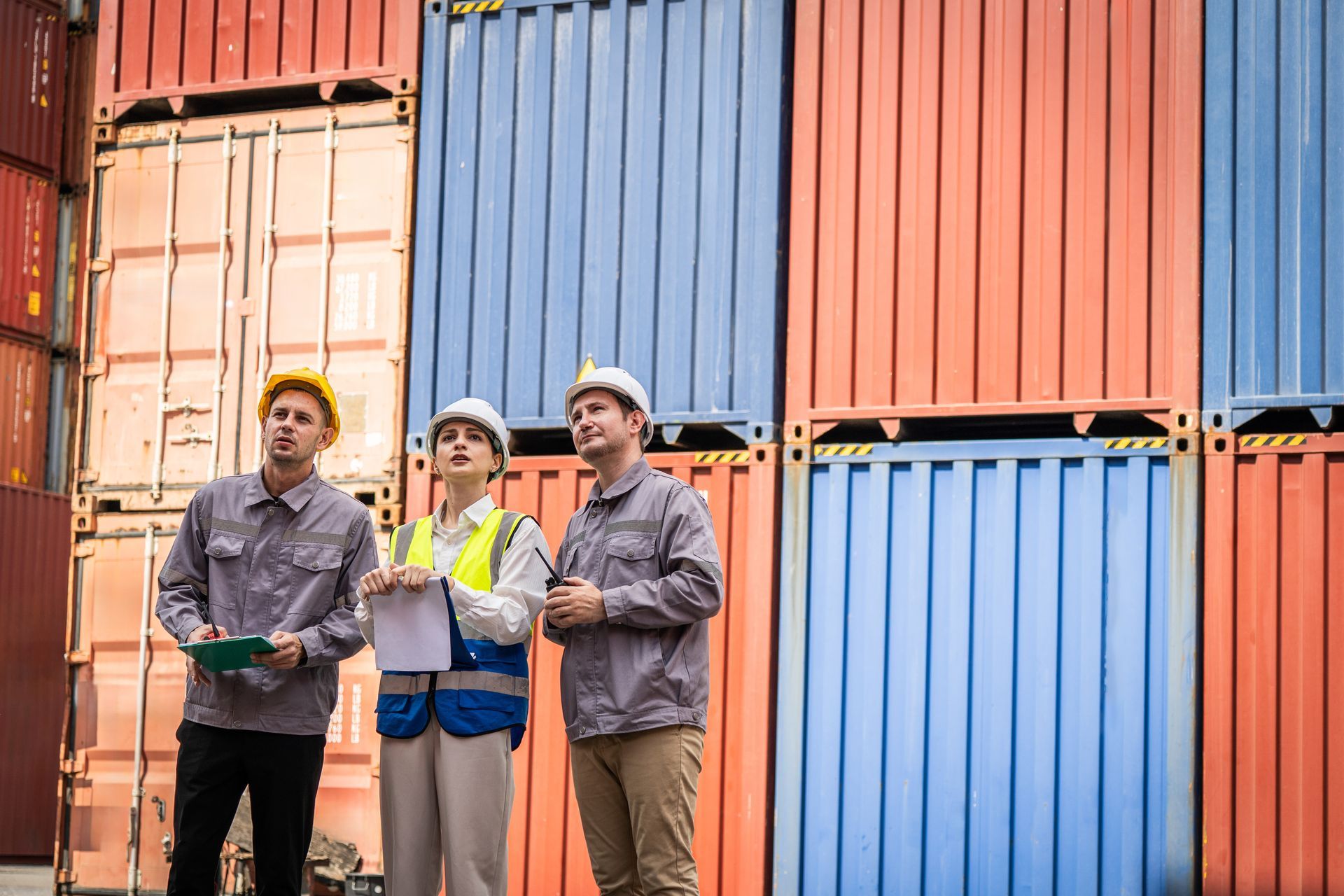 Three workers in hard hats and uniforms stand looking up in front of a tall wall of stacked colorful shipping containers.