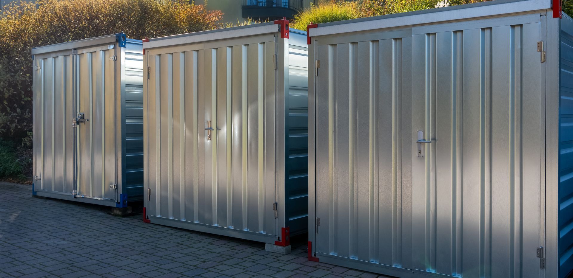 Three metal storage containers in a row, with corrugated sides, on a paved surface, with foliage in the background.