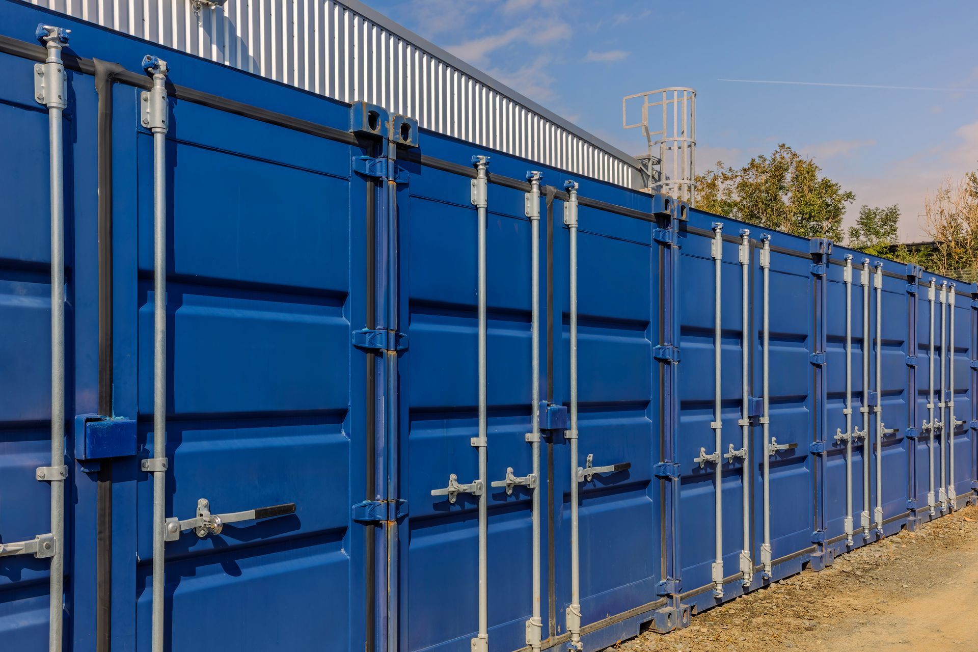 A row of blue shipping containers stands in an outdoor area against a clear blue sky.