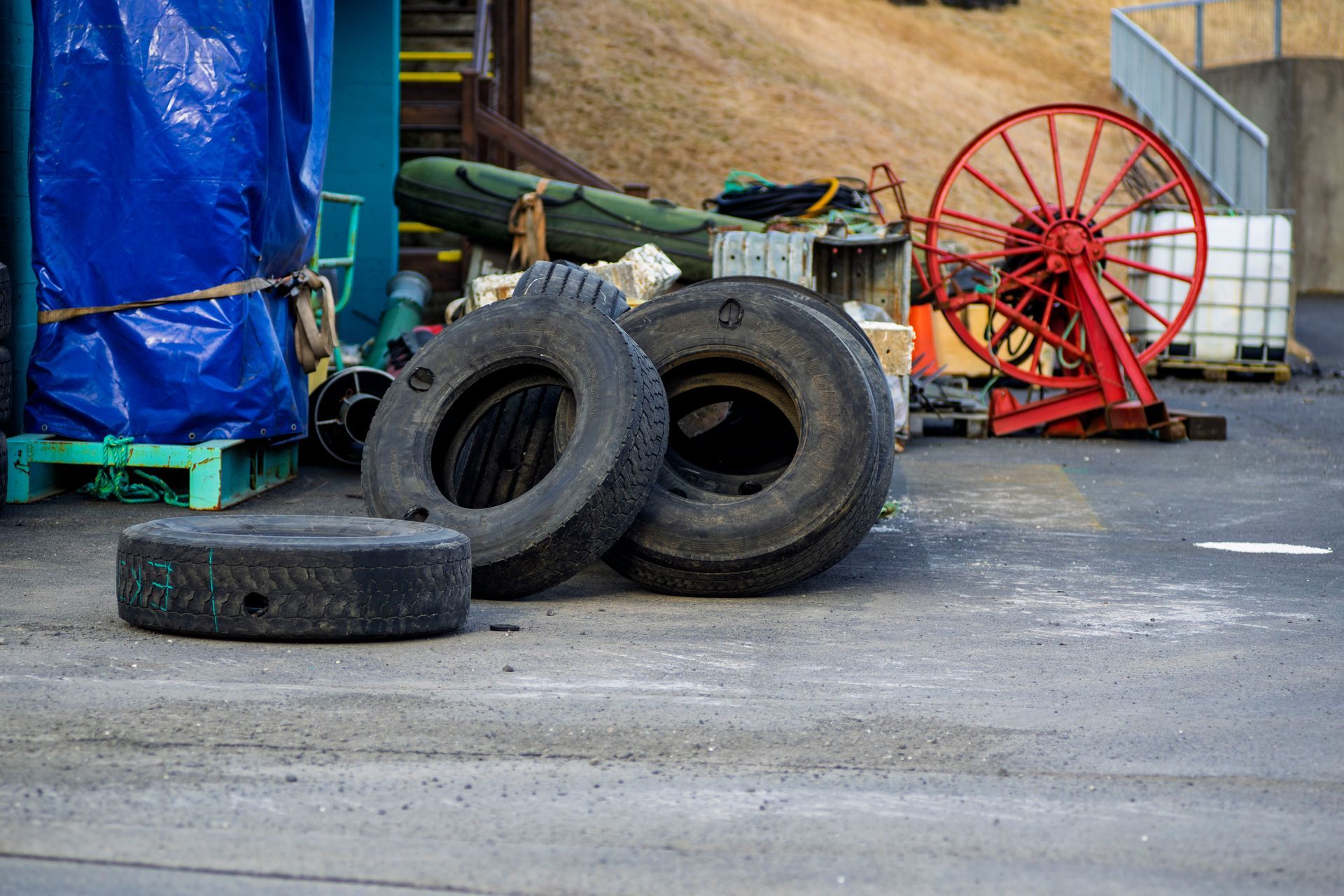 Three worn tires on asphalt; a blue tarp, industrial equipment, and a red fan are in the background.