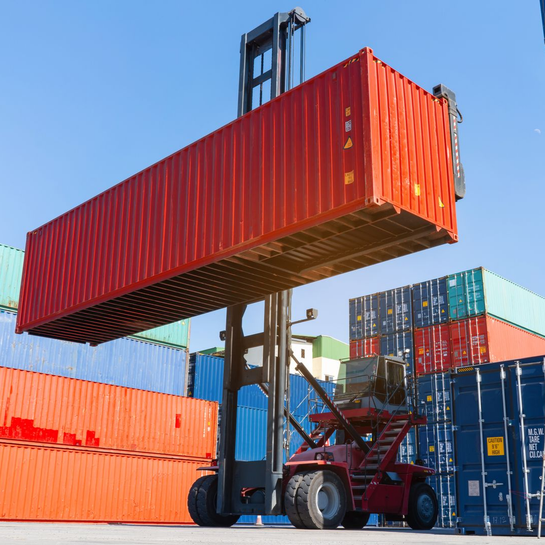 A red reach stacker lifts a large red shipping container high against a clear blue sky in a busy industrial freight yard.