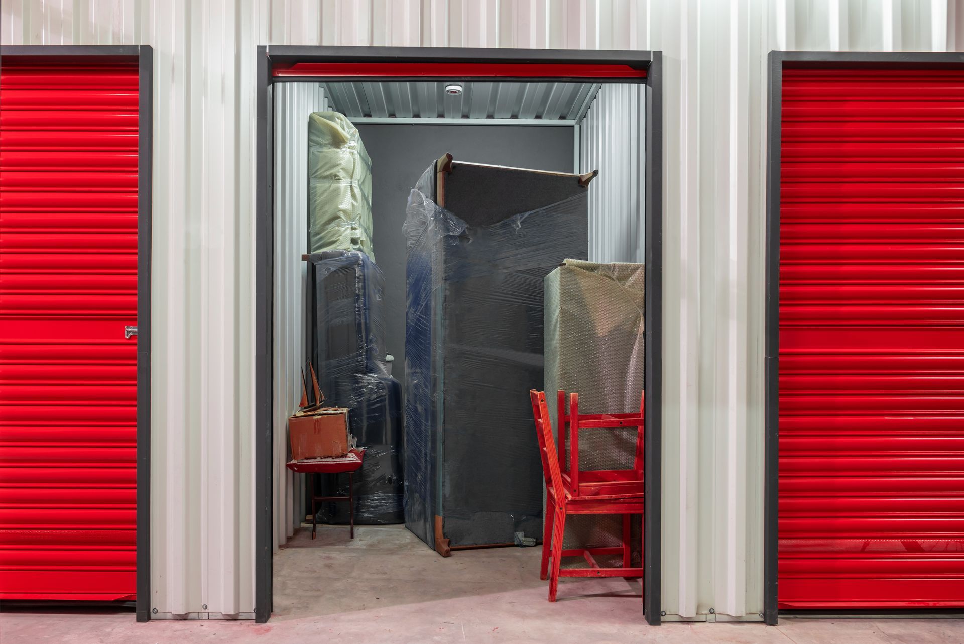 Storage unit with open red door, filled with furniture wrapped in plastic.