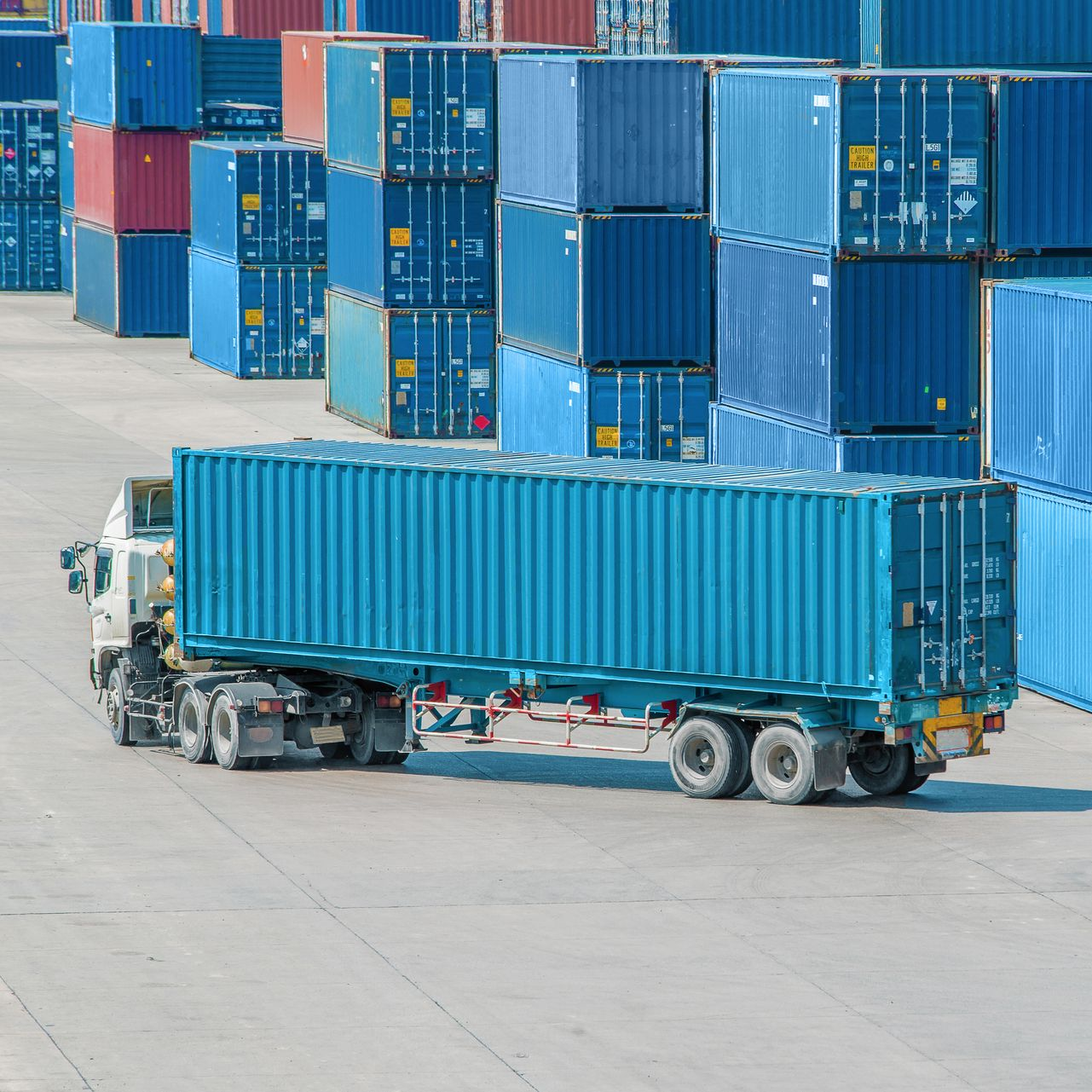 A truck pulling a long blue shipping container across a paved lot filled with stacks of colorful shipping containers.