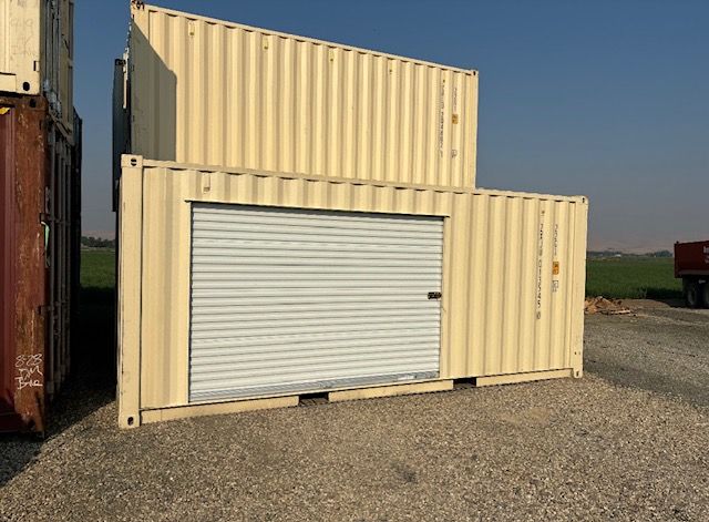 A large, teal, rectangular metal dumpster sits on a paved street next to a sidewalk and parked cars on a sunny day.