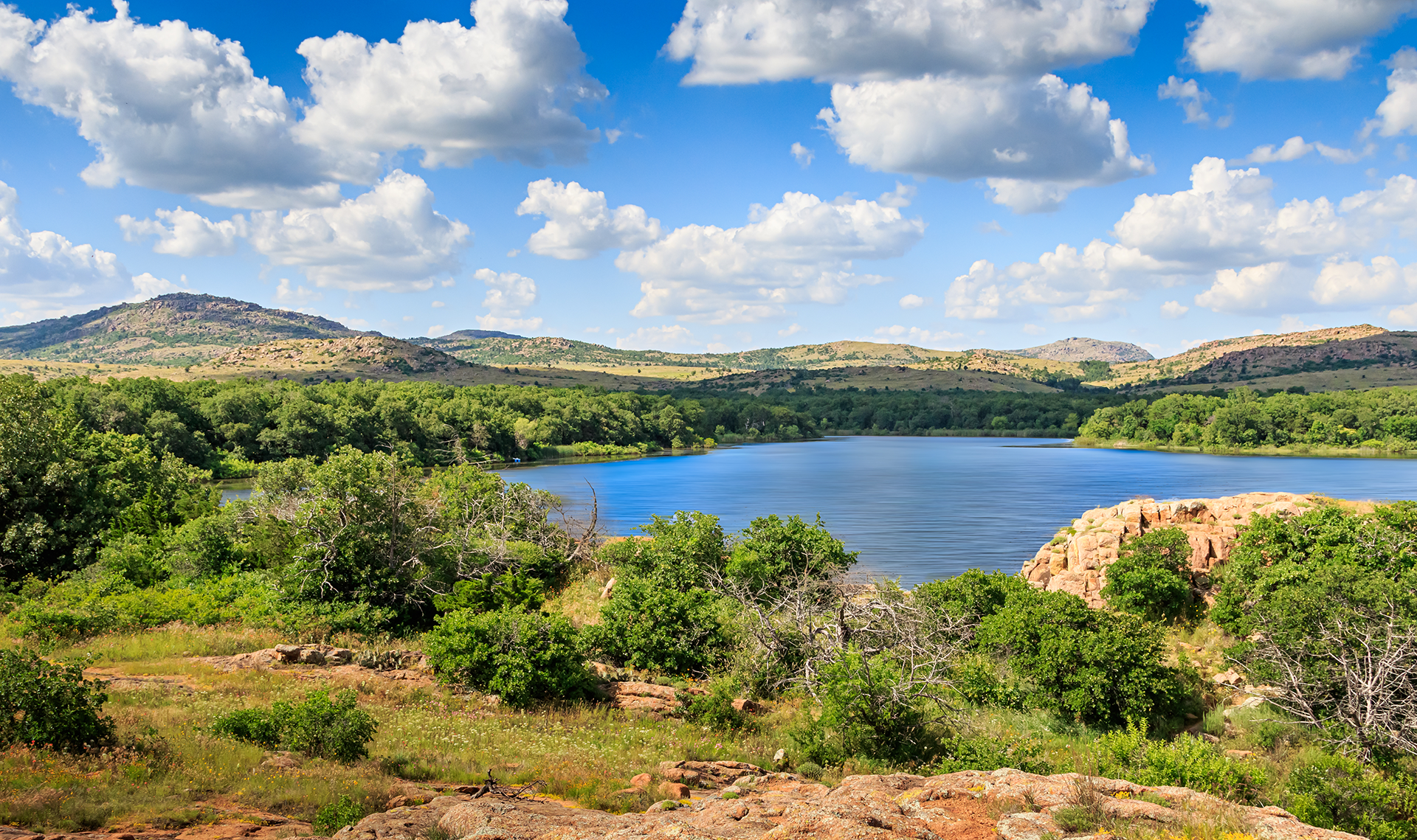 Scenic view of a lake surrounded by green trees and rocky hills under a blue sky with fluffy white clouds.