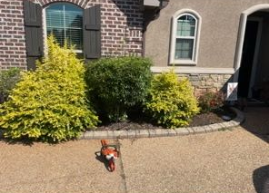 A small, orange toy chainsaw sits on a paved driveway in front of three bushes planted in front of a brick house.
