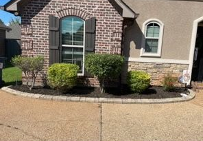A brick and stucco house exterior with a curved stone garden border, dark mulch, and several green shrubs.