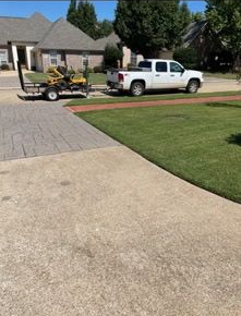 A white pickup truck with a trailer carrying a yellow lawn mower parked on the side of a suburban street.