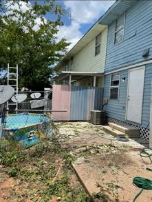 A blue and yellow two-story house with a small pool, patio, and privacy screen in a backyard covered in yard debris.