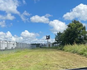 A mowed, grassy field in the foreground, with a line of white trailers behind a fence and a tall billboard in the distance.