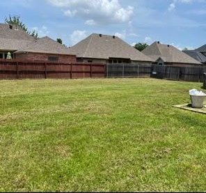 A large, grassy backyard with a wooden privacy fence, brick houses in the background, and a white planter on the right.