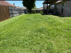 A sunlit residential backyard with a mowed lawn, a wooden fence on the left, and a brick house with a covered patio.