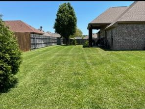 A mowed backyard lawn on a sunny day, featuring a brick house on the right, a wooden fence, and a single tree in the center.