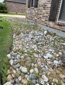 A brick house foundation features a landscaped rock bed overgrown with grass and weeds.