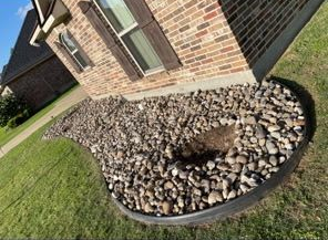 A curved landscaping bed with river rocks against a brick house foundation, featuring a small empty dirt patch.