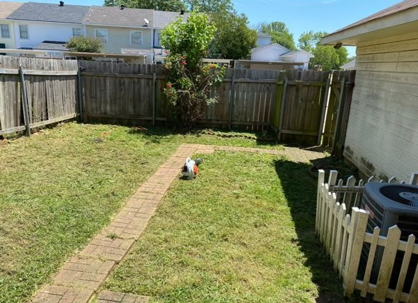 A fenced-in backyard with a brick path, a lawnmower in the center, and an air conditioning unit behind a small white fence.