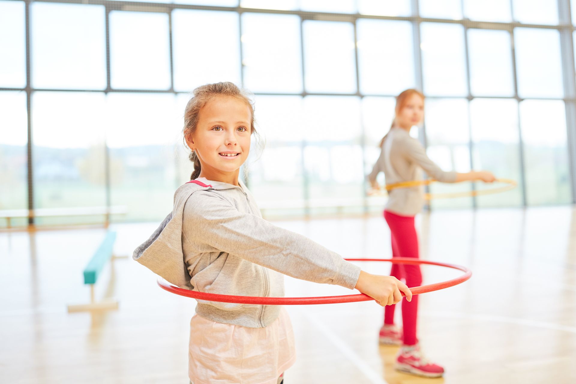 Zwei junge Mädchen spielen in einer Turnhalle mit Hula-Hoop-Reifen.