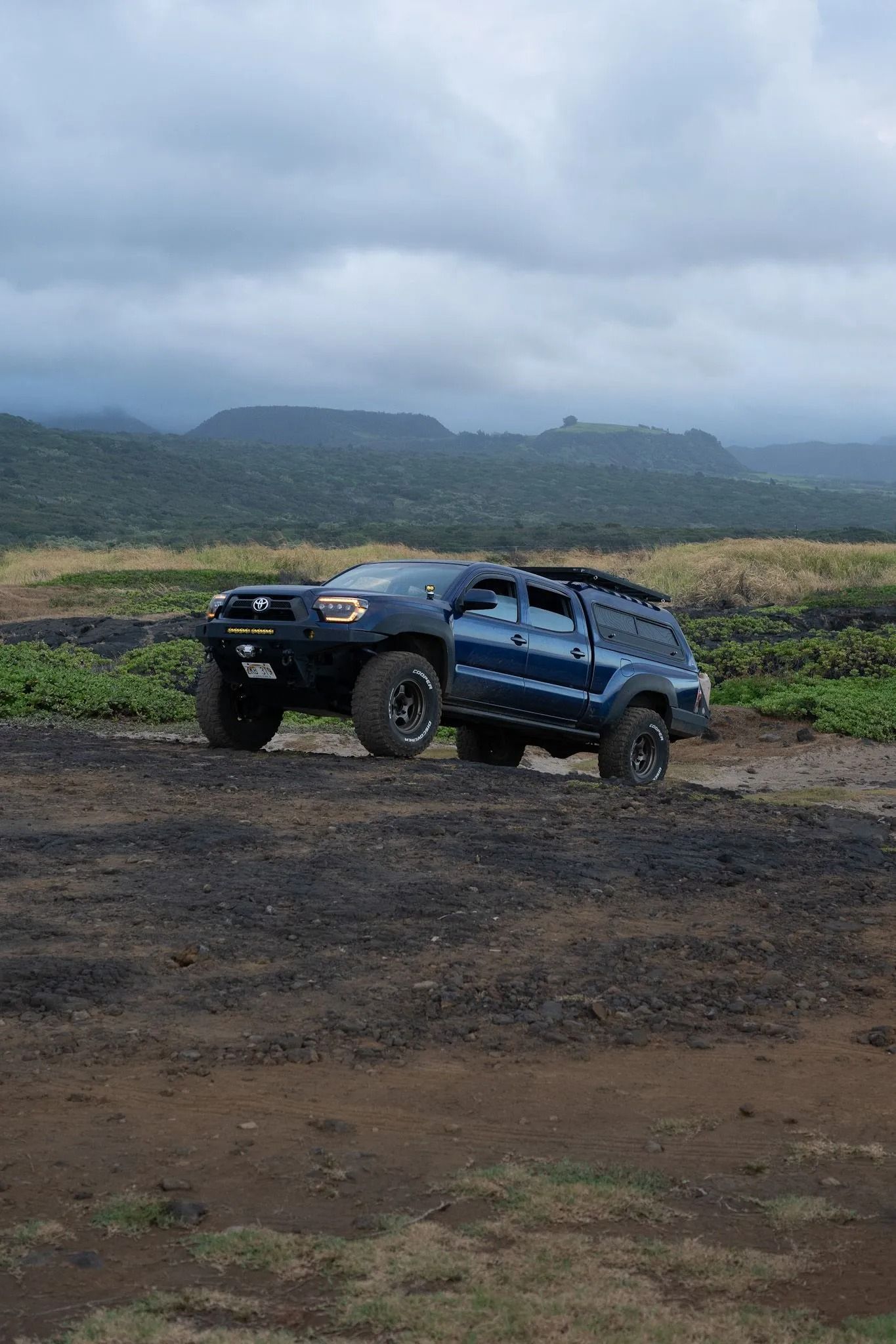 A blue truck is parked in a dirt field with mountains in the background.