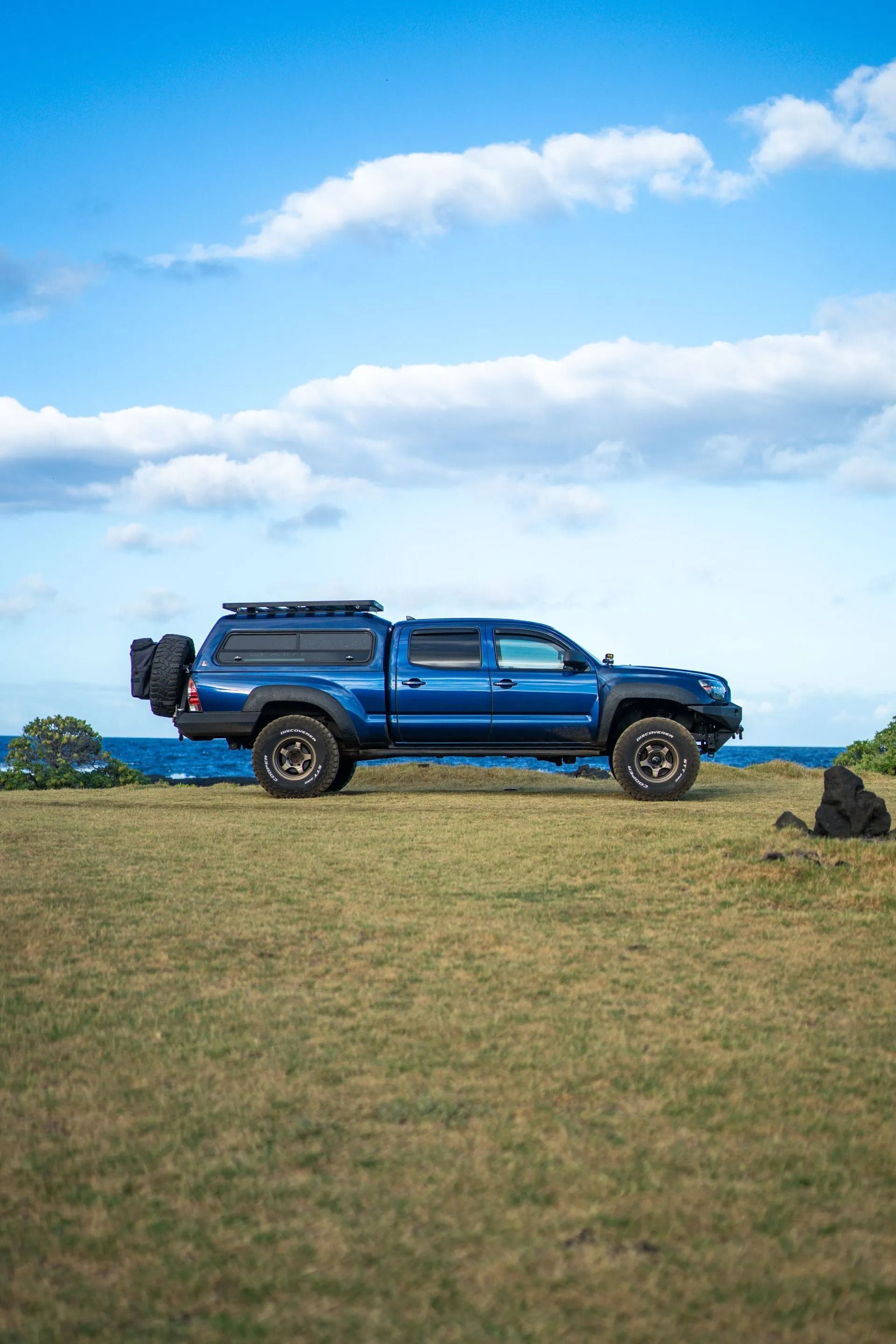 A blue truck is parked in a grassy field near the ocean.
