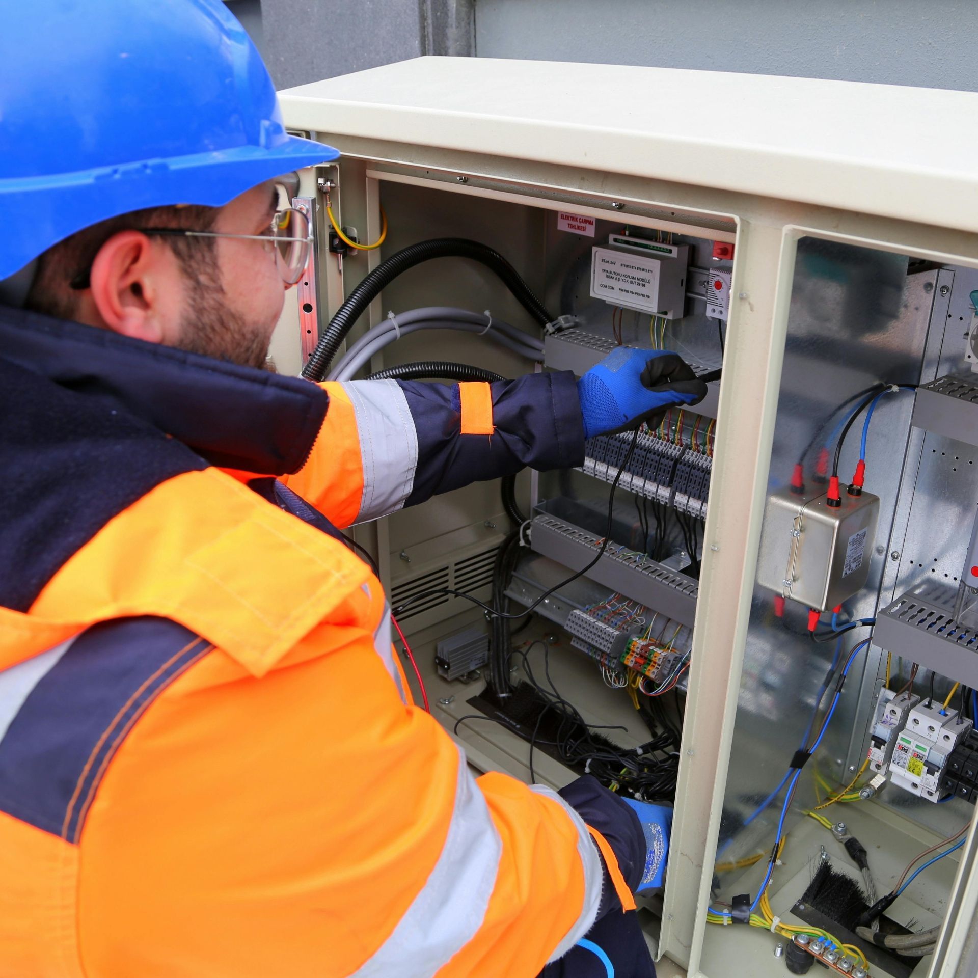 MJS Electrical Services - Electrician in blue hard hat, orange jacket, gloves, working on a panel in an outdoor setting.