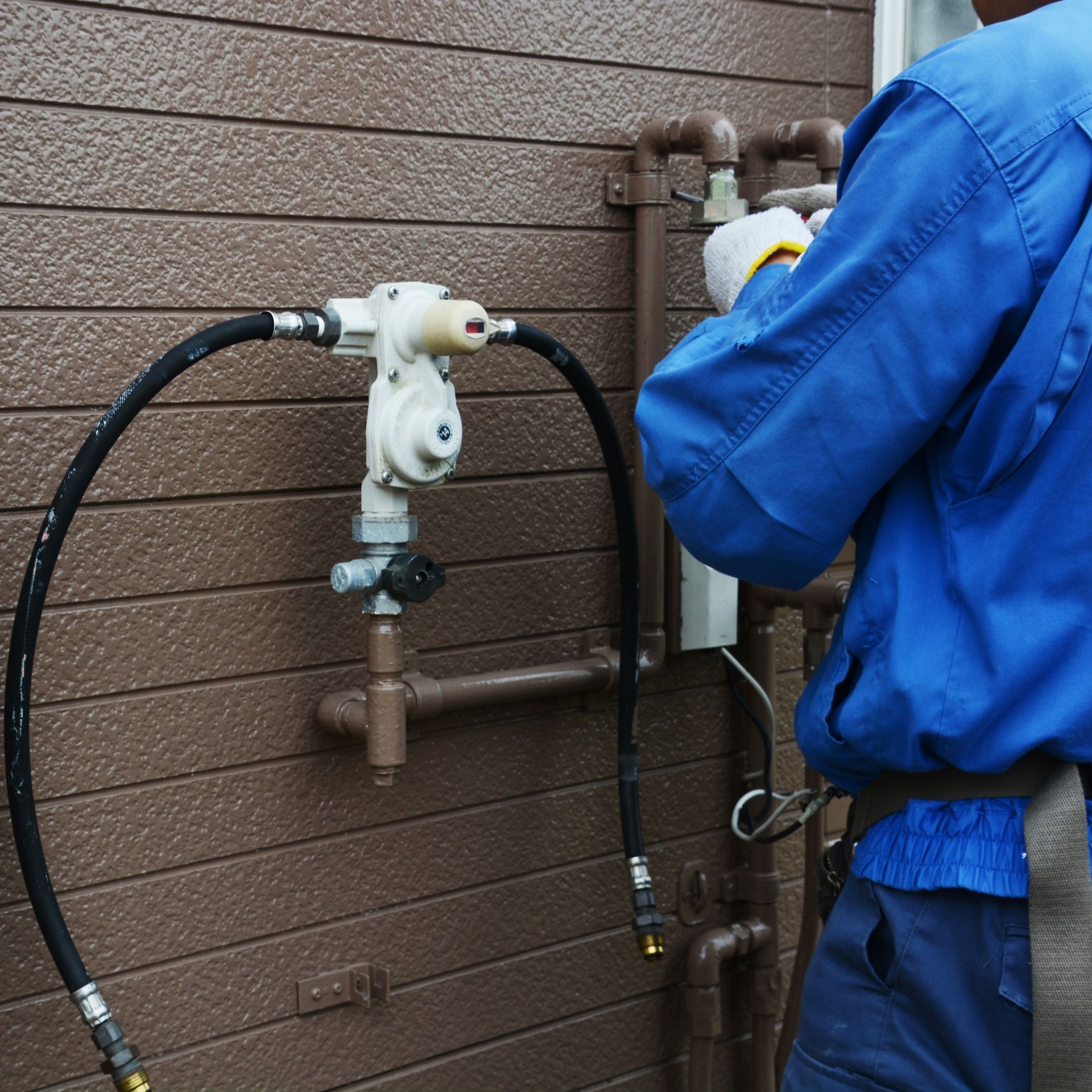 MJS Electrical Services - Plumber in blue overalls works on gas line outside brown building.