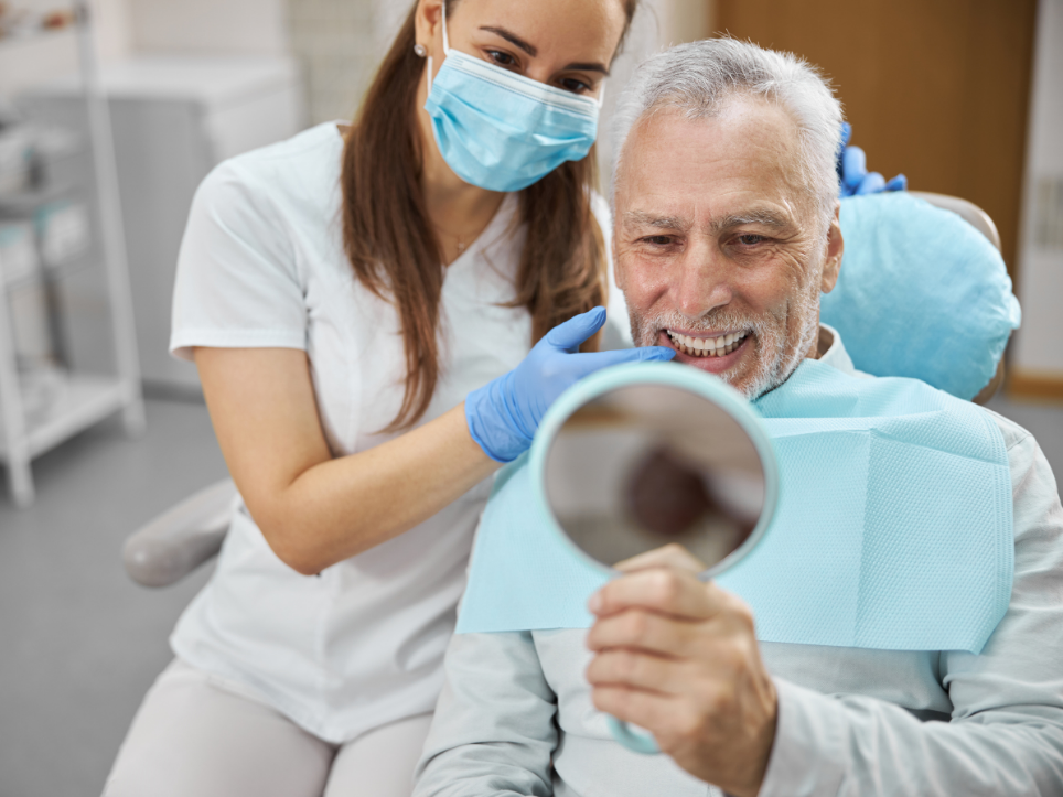 A photo of a senior man looking at his new smile in a mirror with a dental assistant