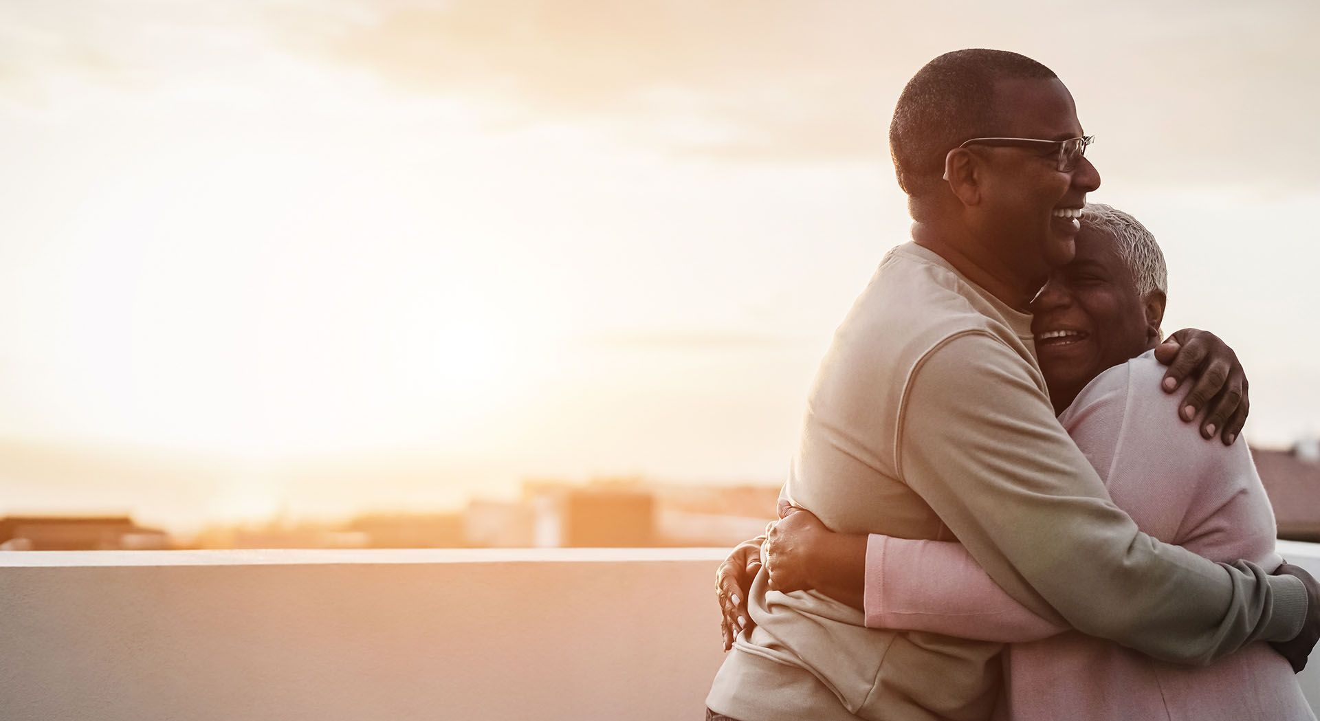 An elderly couple hugging each other on a balcony at sunset.