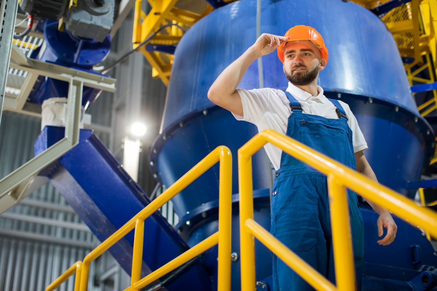 Trabajador con mono azul y casco naranja de pie sobre una plataforma industrial cerca de maquinaria de gran tamaño.