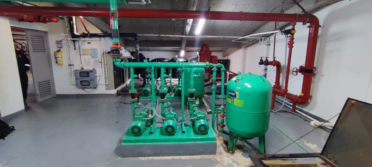 Sistema industrial de bombeo y tuberías en un cuarto de servicio, con tuberías rojas en el techo y un tanque de presión verde.