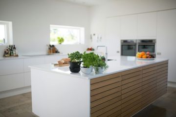 Modern white kitchen with island, featuring plants and fruit on a light countertop and wood-paneled island base.