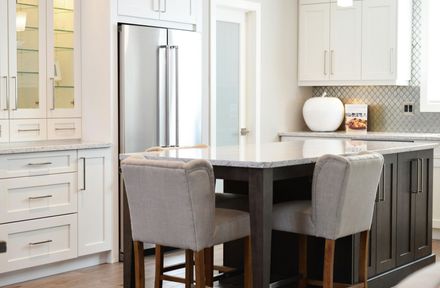 Modern kitchen with island and two gray upholstered bar stools. White cabinets and stainless steel refrigerator.