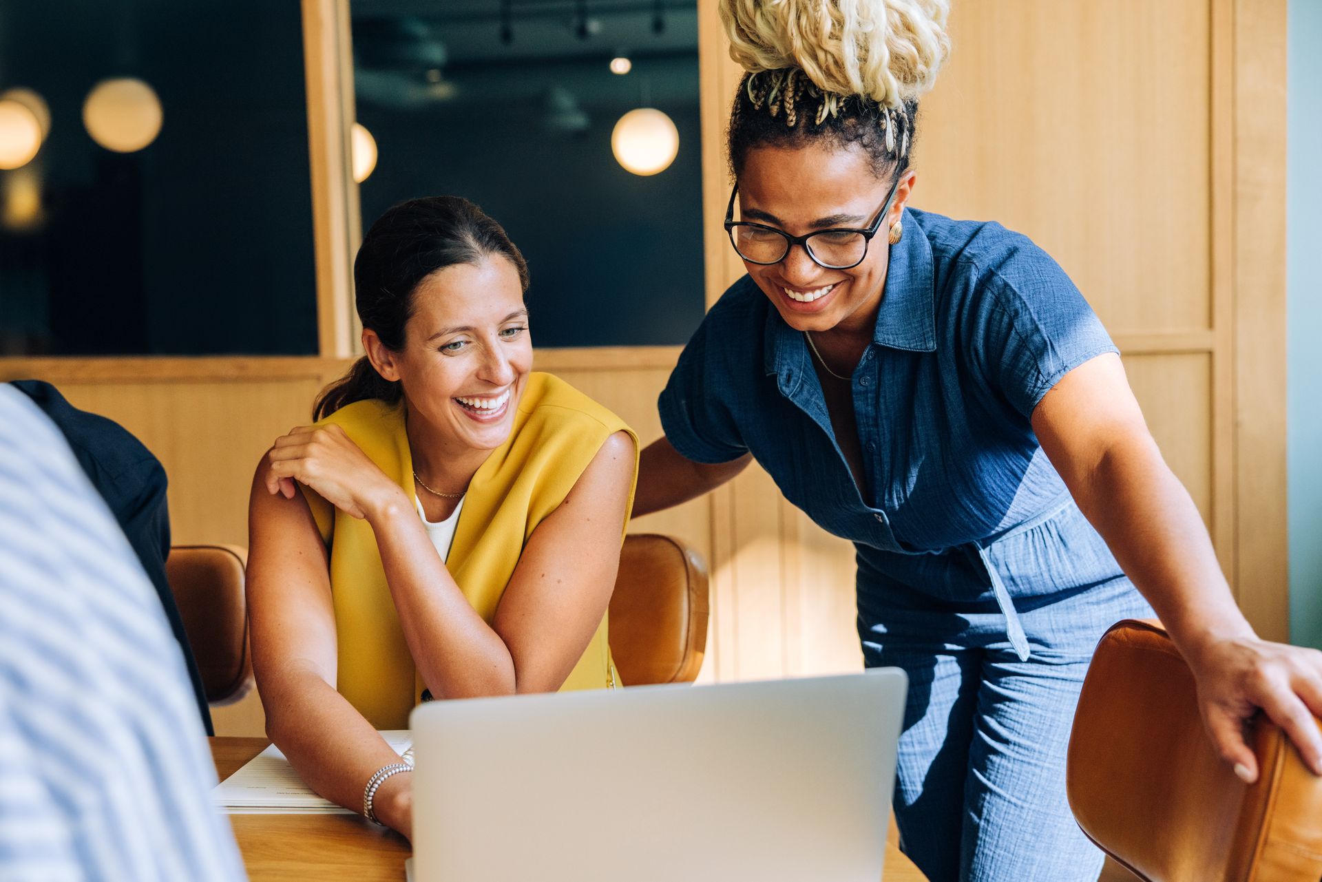 Two women smiling, looking at a laptop in a brightly lit office setting. One has glasses and light-colored hair.