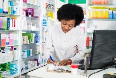 Pharmacist at a pharmacy counter, writing, with shelves of medication in the background.