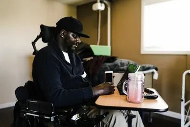 Man in wheelchair using phone and tablet at a table; drink nearby. Indoor setting.