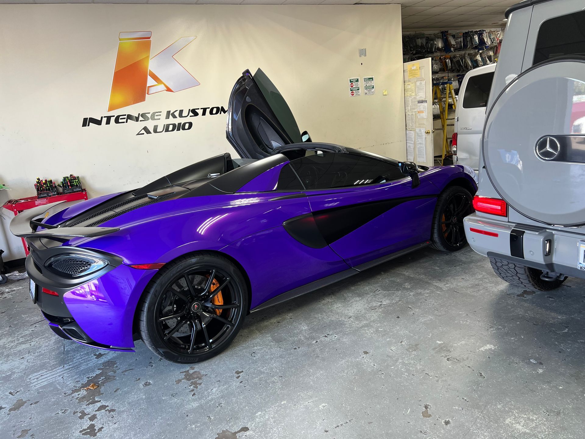Purple McLaren with doors open, parked in a garage next to a Mercedes.