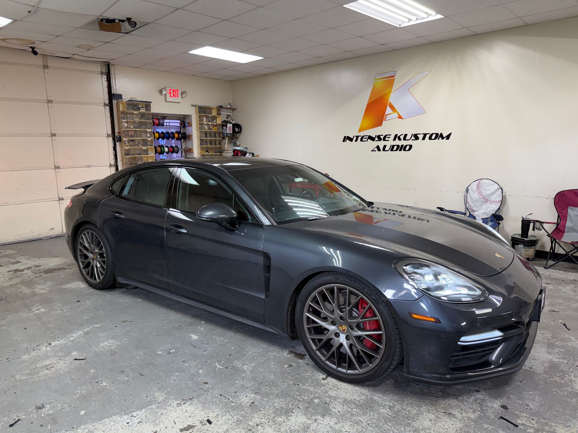 Gray Porsche Panamera in an audio shop with red brake calipers, logo on wall.